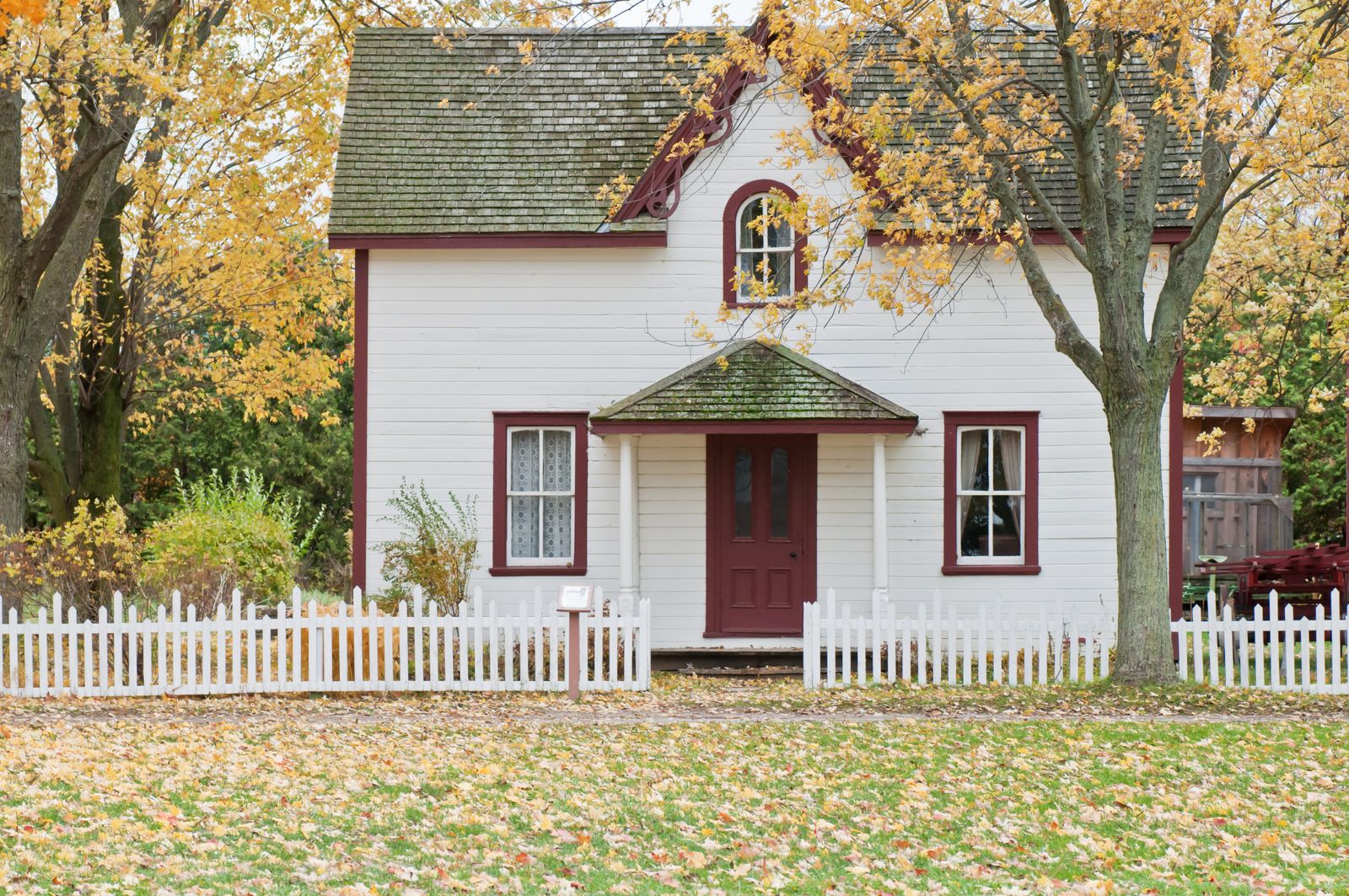 A classic white picket fence in front of a home with autumn trees